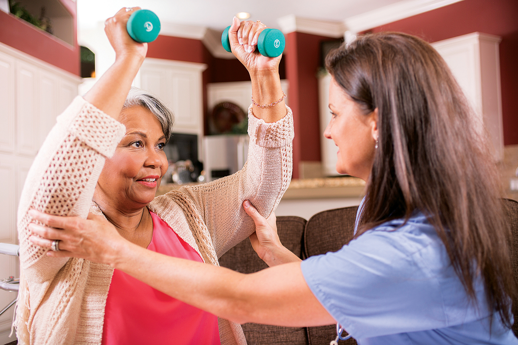 Caring, Latin descent home healthcare nurse conducts physical therapy exercises with African descent senior adult patient at home, assisted living, or nursing home setting. She helps her with arm strengthening exercises using a dumbbell. Kitchen background.