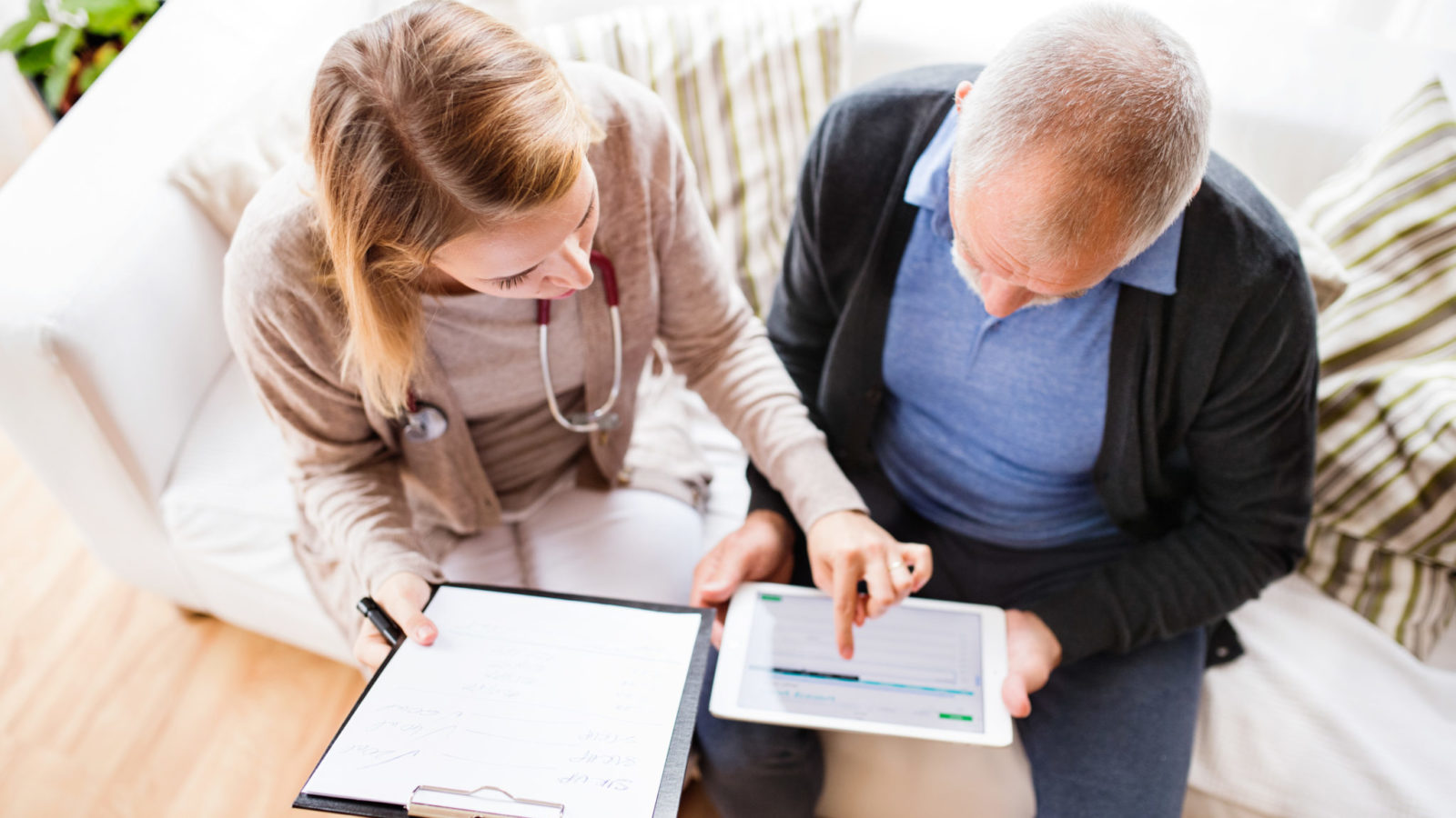 Health visitor and a senior man during home visit. A female nurse or a doctor showing test results on a tablet. High angle view.