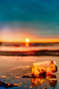 a mason jar with string lights lays on the ocean floor with waves approaching it as the sun sets in the distance