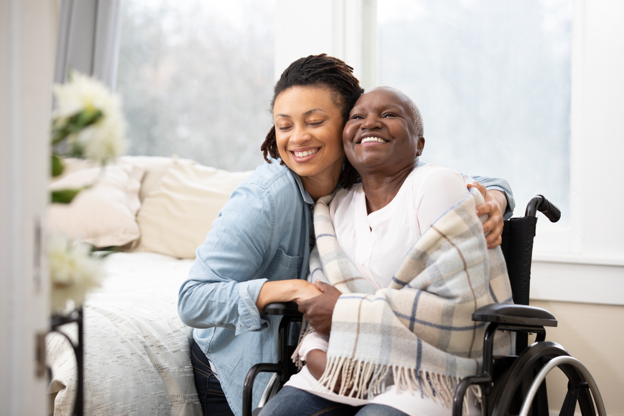 A daughter puts her arms around a smiling mother wrapped up in a blanket, sitting in a wheelchair.