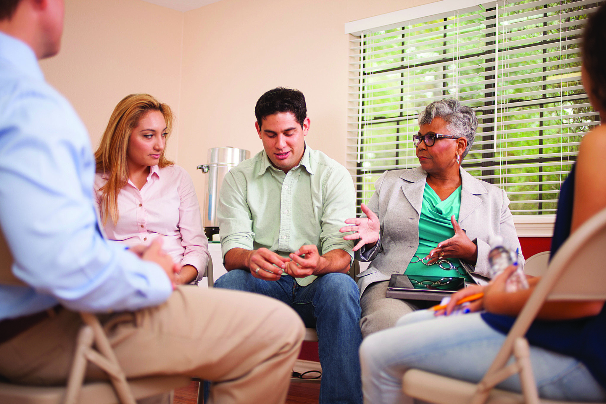 Multi-ethnic, mixed age group of people in a group counseling session with a therapist. The female counselor, psychiatrist, or therapist is taking notes on her digital tablet. The rest of the group is talking, sharing together in an office setting.