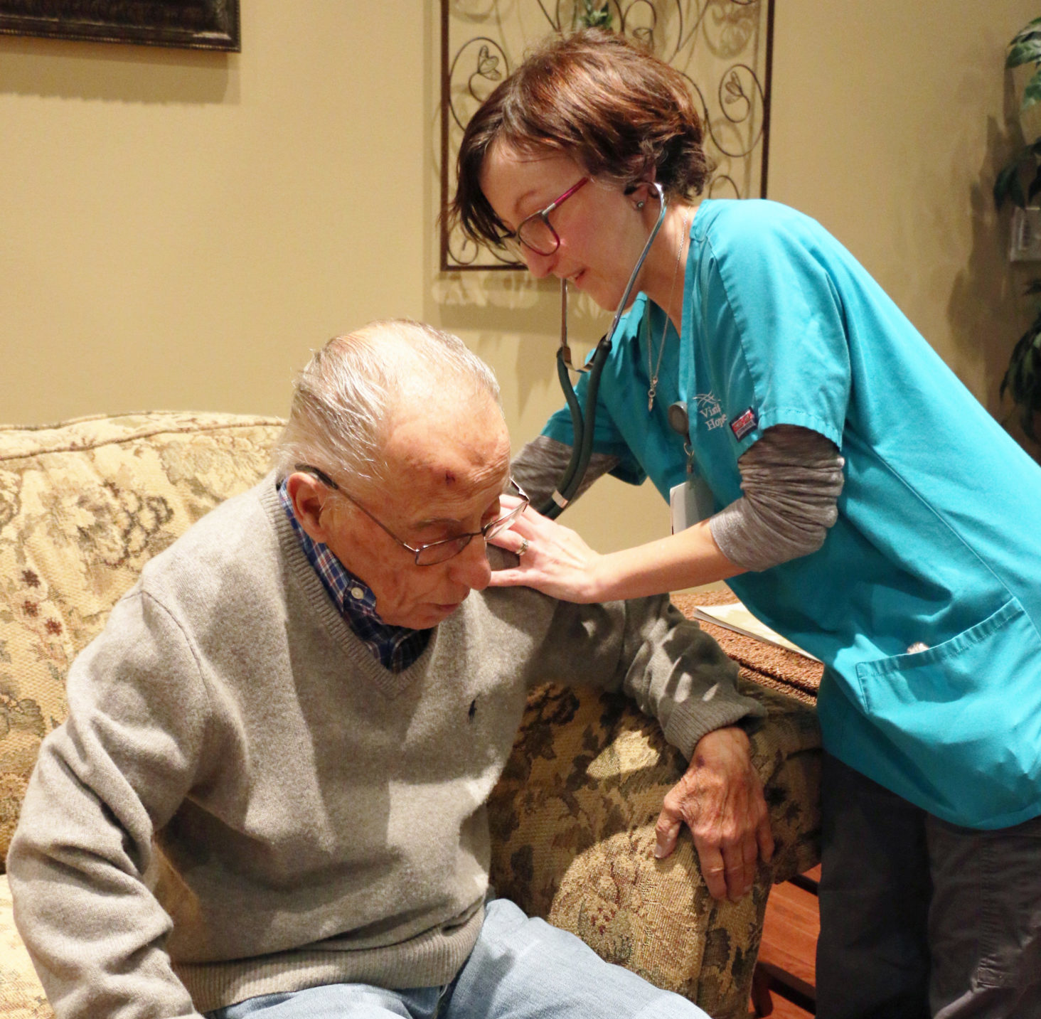 Young clinician with short hair and glasses checks the heart rate of a sitting older gentleman