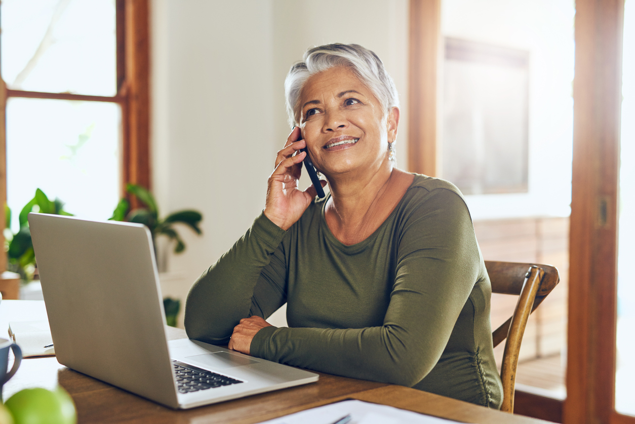 Shot of a mature woman talking on a cellphone while working on a laptop at home