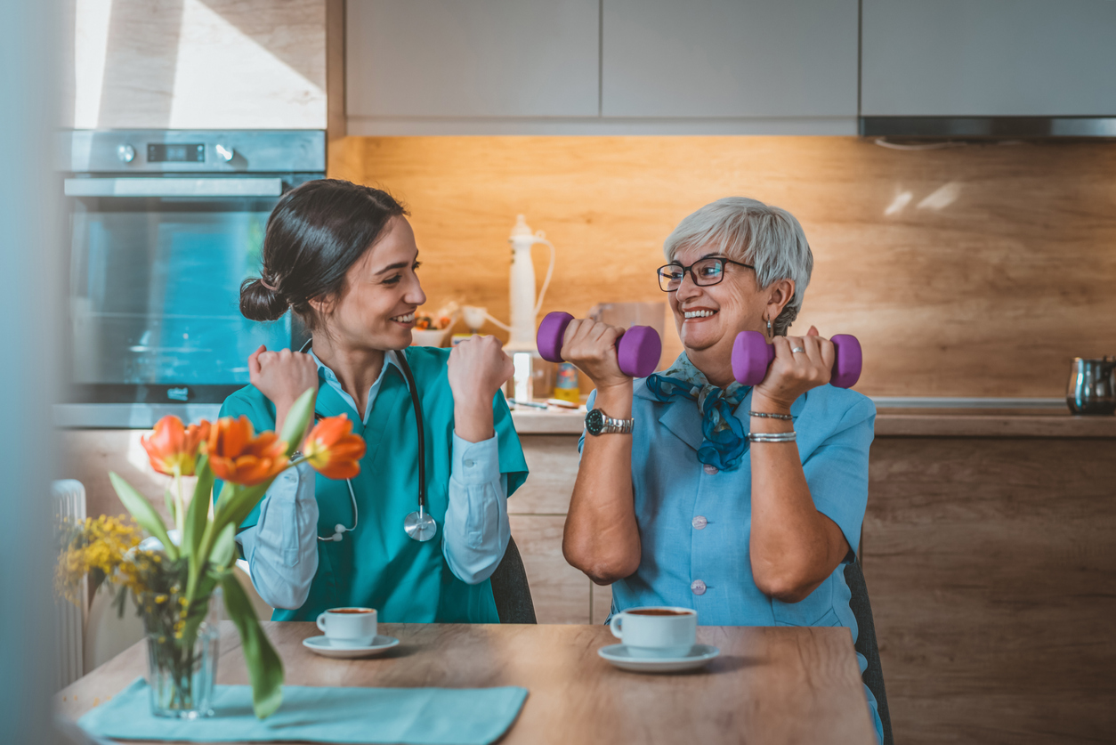 Photo of Nurse helping senior woman in lifting dumbbell at retirement home during the day. Senior woman with dumbbells in rehab with a physiotherapist smiling. Physiotherapist helps elderly woman with healthy dumbbell training in rehab or home in the kitchen while sitting at table.