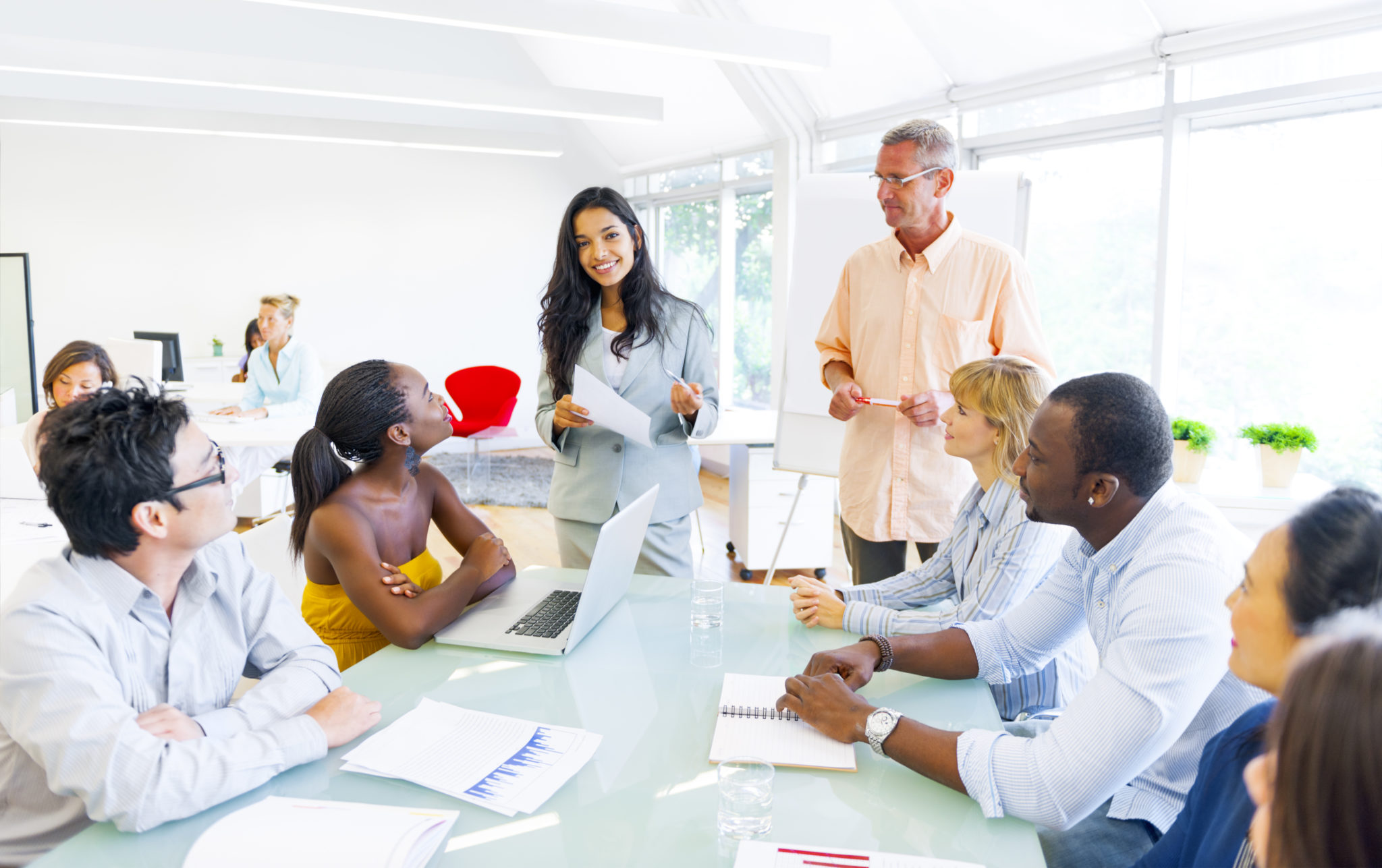 Business Training Woman in a business suit smiles at the camera as a table full of people look up at her