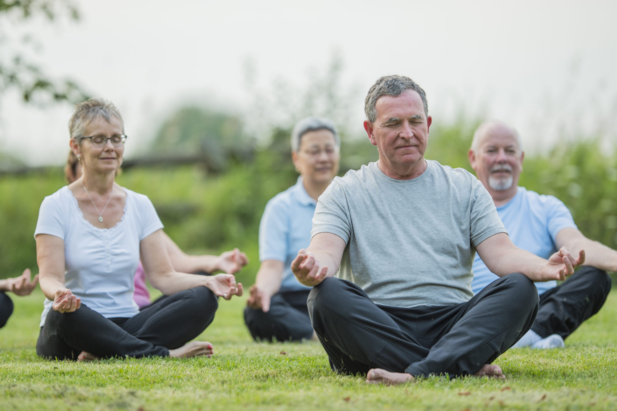 A multi-ethnic group of senior adults are taking a tai chi fitness class. They are practicing their martial art together in a park at sunset. They are enjoying the sunlight and summer weather. Here they take a break to meditate.