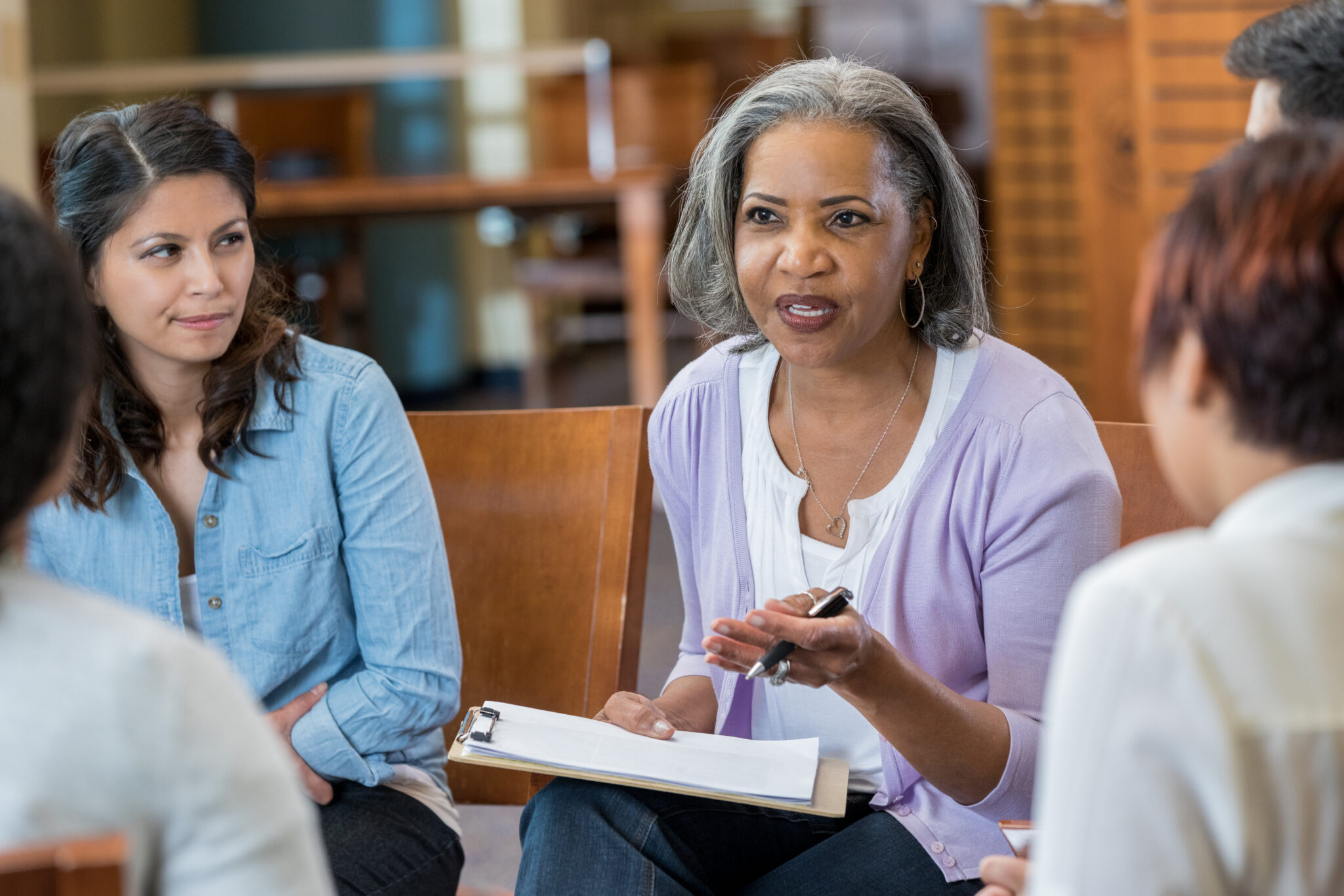 Senior mental health professional facilitates support group Serious senior African American female mental health profession gestures while talking with clients in a support group or group therapy session. She is holding a clipboard.