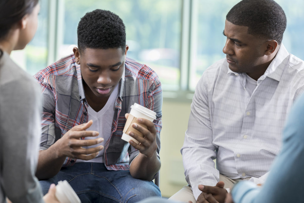 Vulnerable man in support group Young African American man has his head down while talking about issues during a group therapy session. A concerned therapist is sitting next to him.