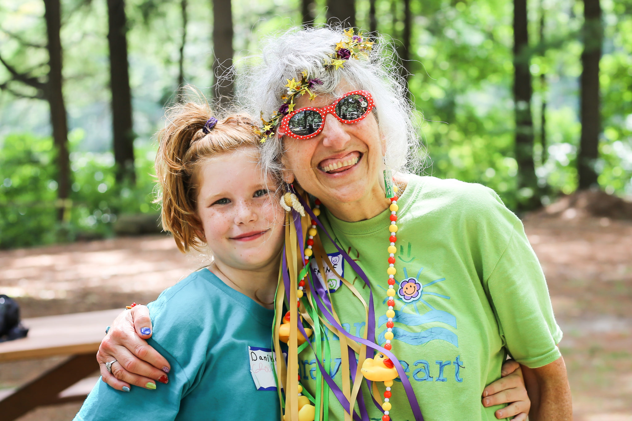 Smiling at Camp BraveHeart young girl with red hair poses next to an older woman who is wearing red polka dot sunglasses