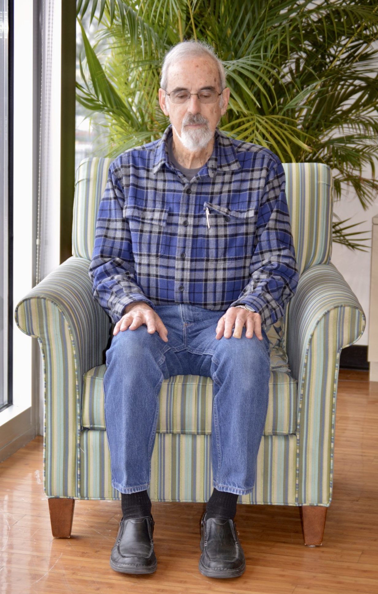 Older man with glasses on his face sits in a green stripe chair with his hands on his knees. A vibrant green plant is in the background.