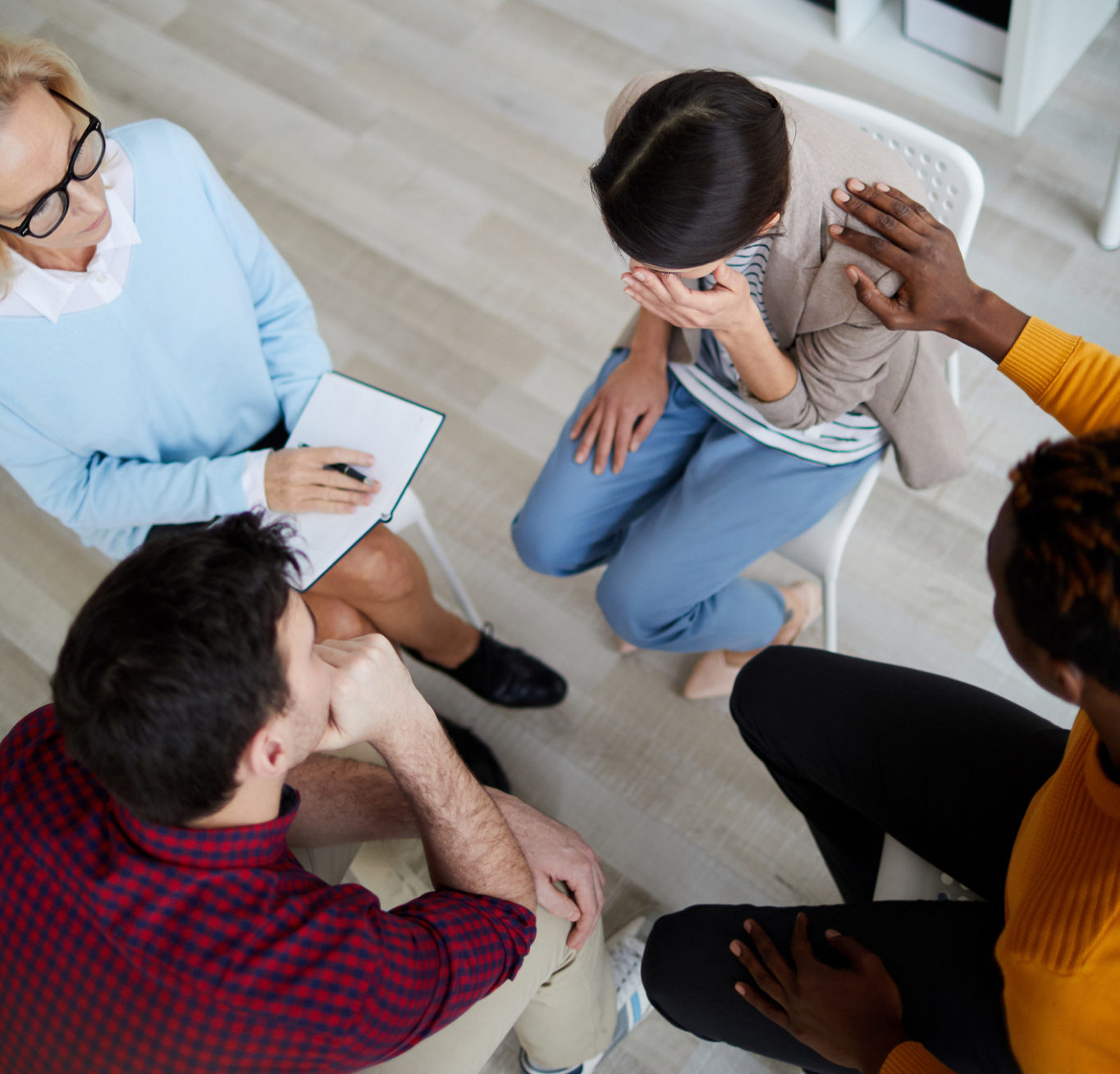 Directly above view of support group sitting in circle at therapy session and discussing problems of each other, black man consoling crying woman