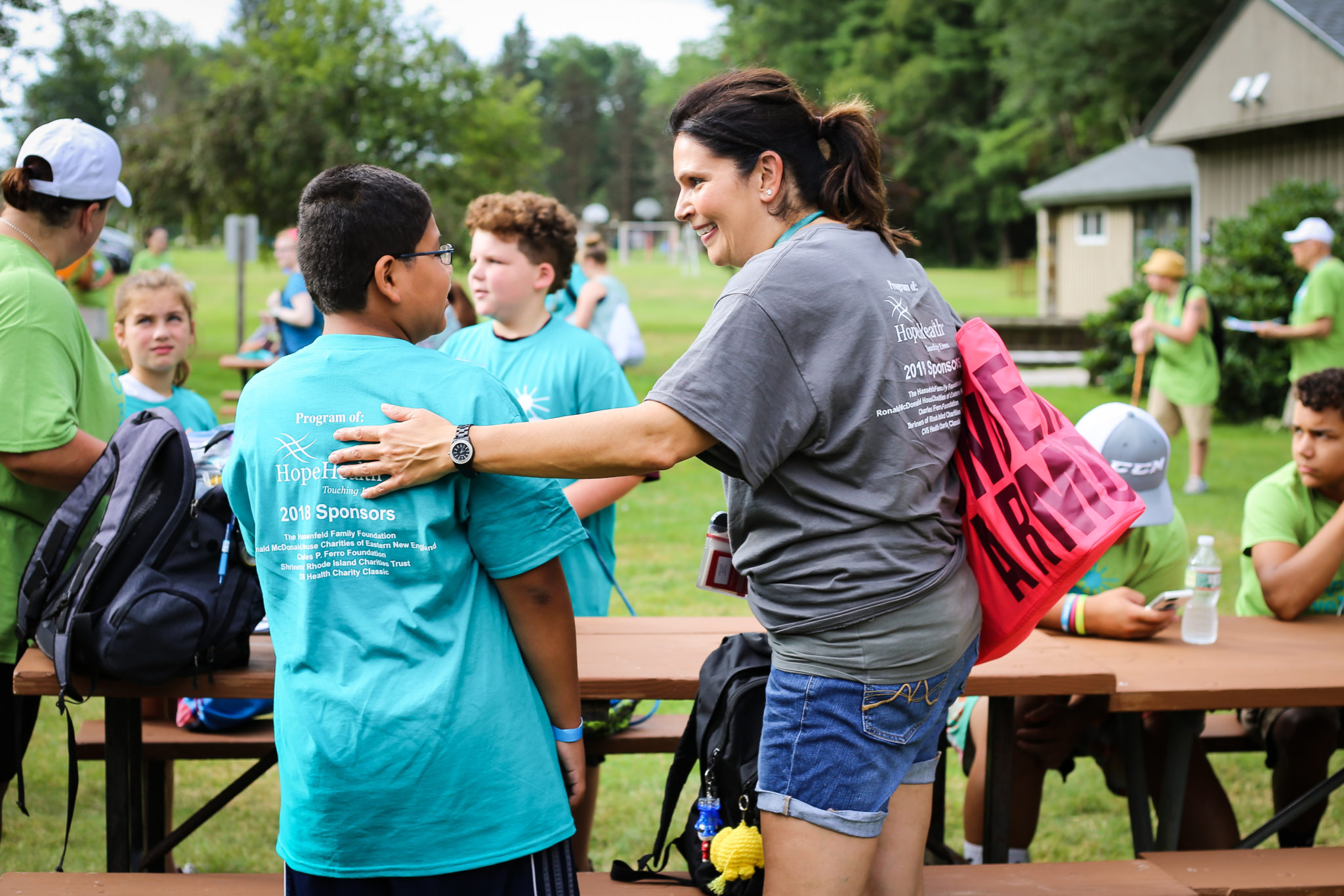 IMG_8214 A woman in a grey camp shirt places her had commpasionately on the shoulder of a boy wearing a teal camp shirt. Camp kids are in the background of the scene.