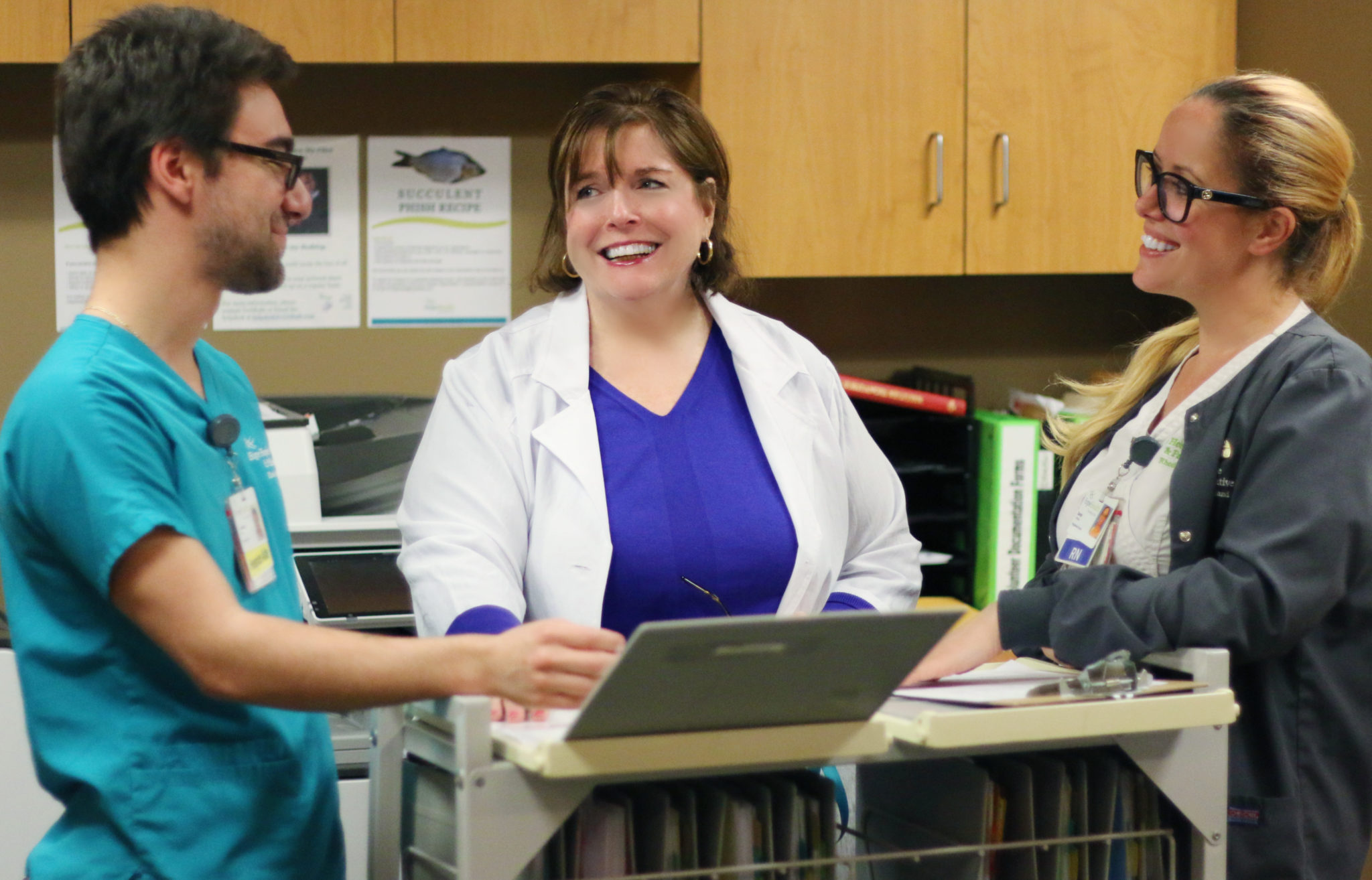 Young male in teal scrubs talking to two female nurses at the nurses station while smiling