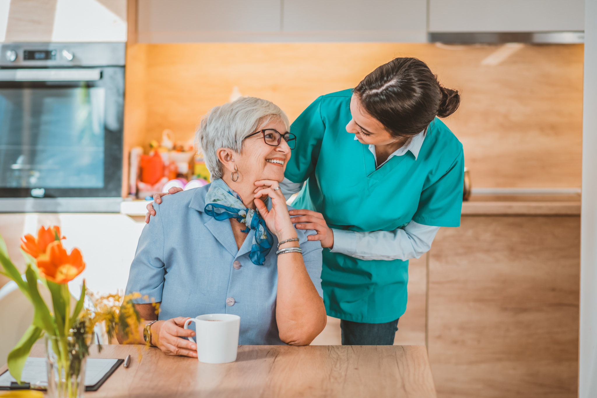 Photo of young female Health visitor and a senior woman during home visit. Helpful young pretty woman and lovely older lady. Home caregiver standing and senior woman sitting at nursing home. Female nurse consulting with elderly patient, looking at each other with smile.