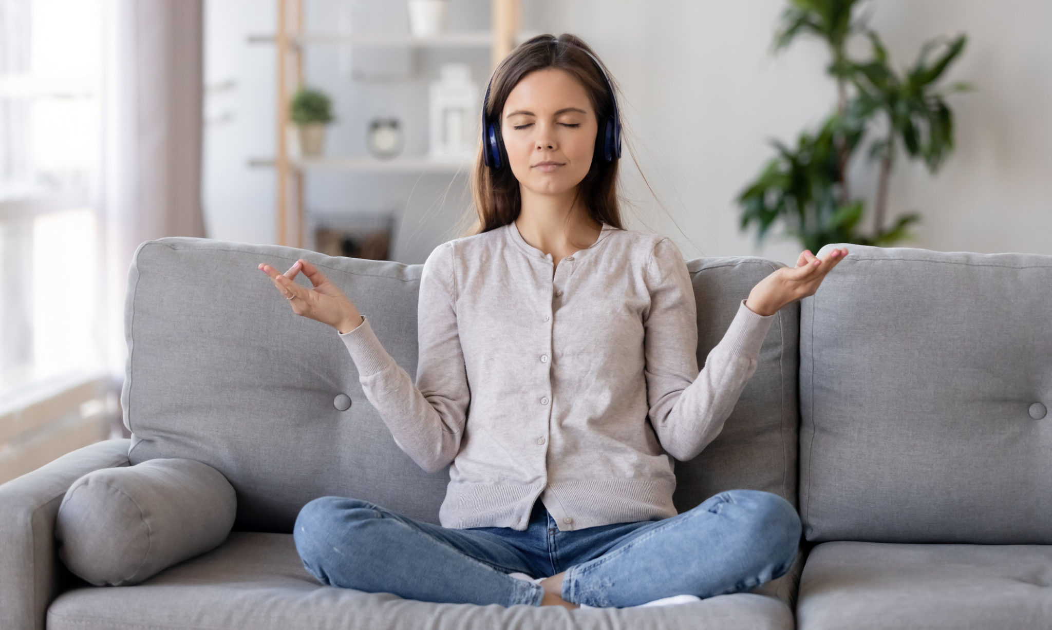 Calm young woman sit on couch meditate with mudra hands listen to slow music in headphones, peaceful relaxed girl female wearing earphones practice yoga on sofa enjoy rhythmic chillout tracks