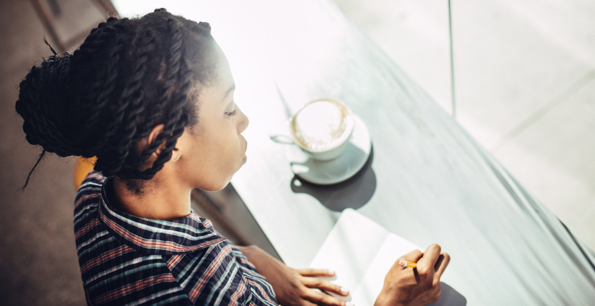 A beautiful young African American woman sits at the window counter of a coffee shop, enjoying a latte while writing ideas down in a small notepad journal or diary. She wears a casual button up shirt, with her hair up, and a content relaxed look on her face. Shot in downtown Los Angeles. Bright sunlight cascades in through the windows illuminating the pages of paper and hot drink. Horizontal with copy space.