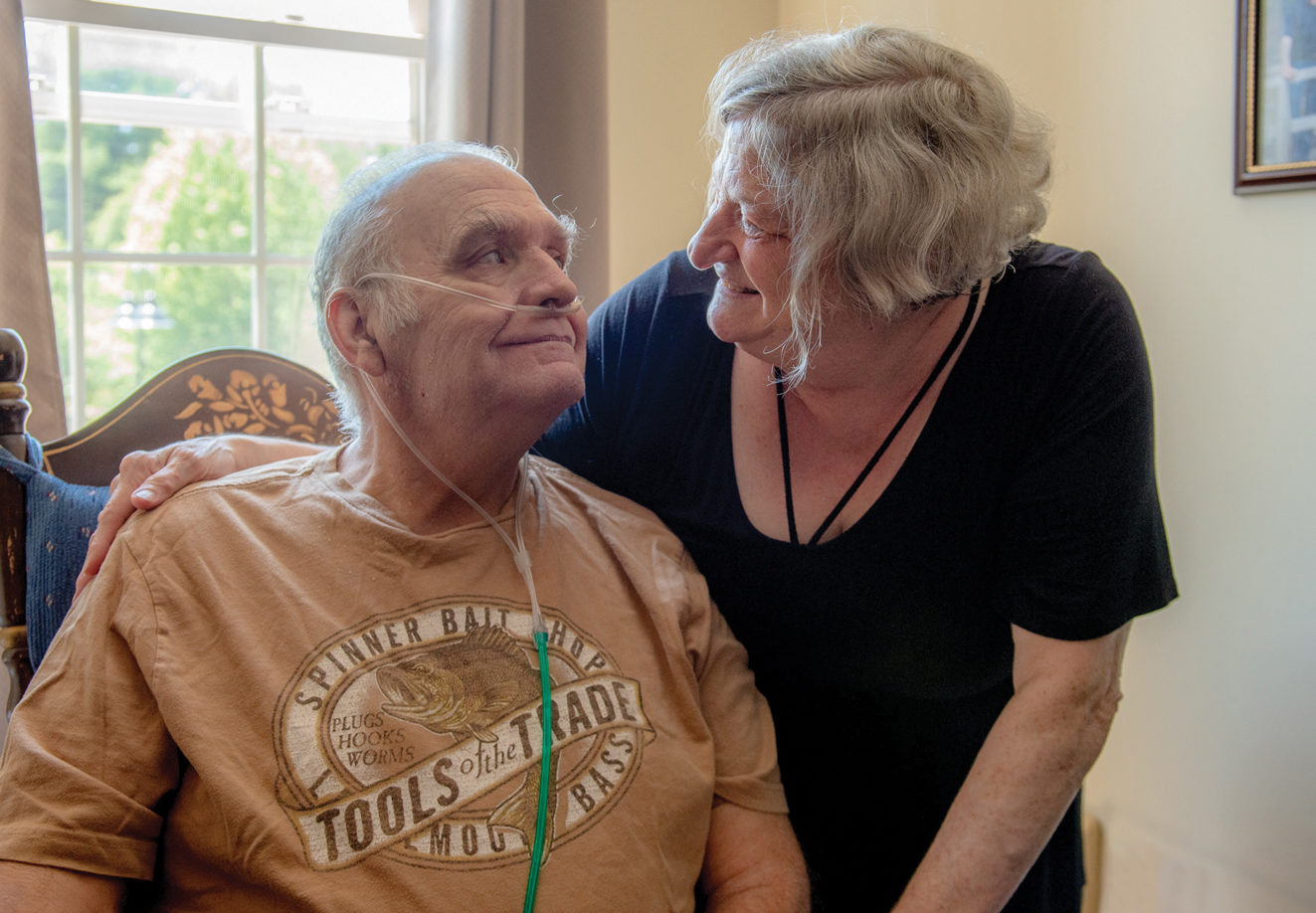 An elderly couple looks lovingly at each other. The man is sitting and wearing an oxygen tank while the woman is standing next to him with her arm around him.