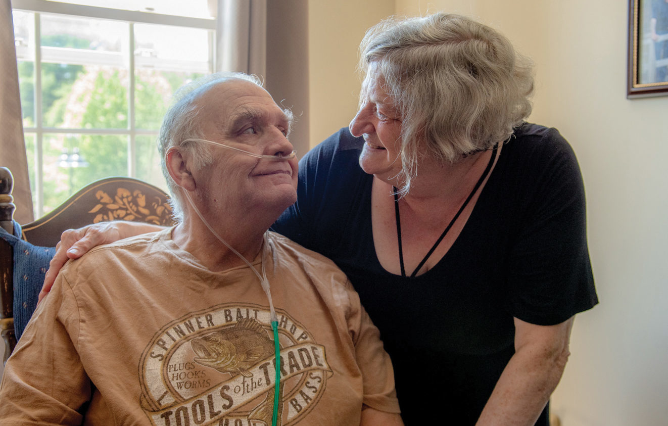 An elderly couple looks lovingly at each other. The man is sitting and wearing an oxygen tank while the woman is standing next to him with her arm around him.