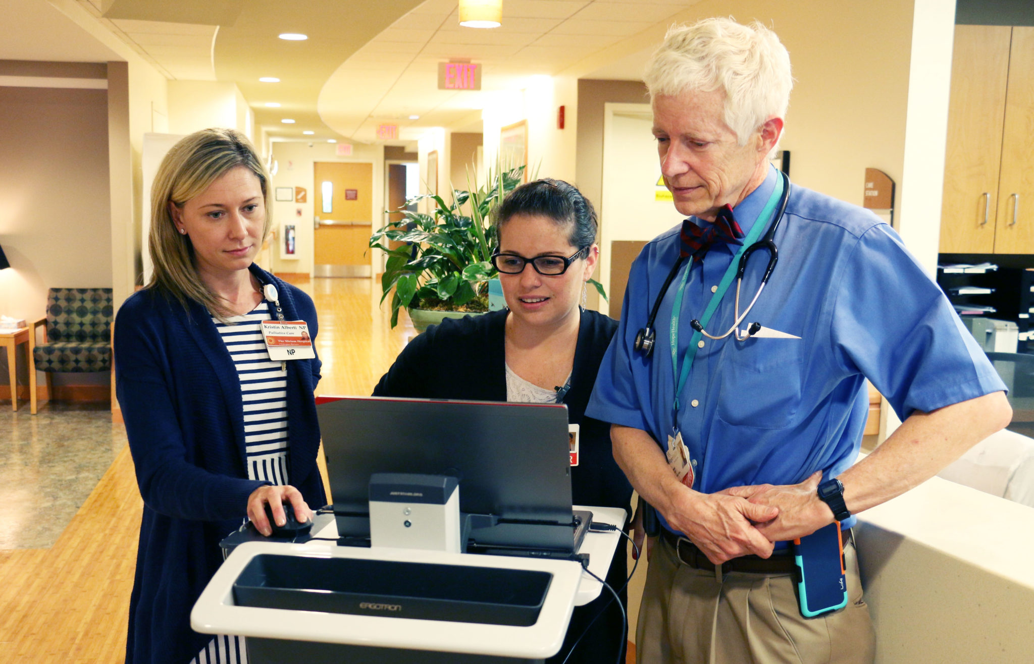 Two young women stand with an older man while reviewing a laptop in front of them