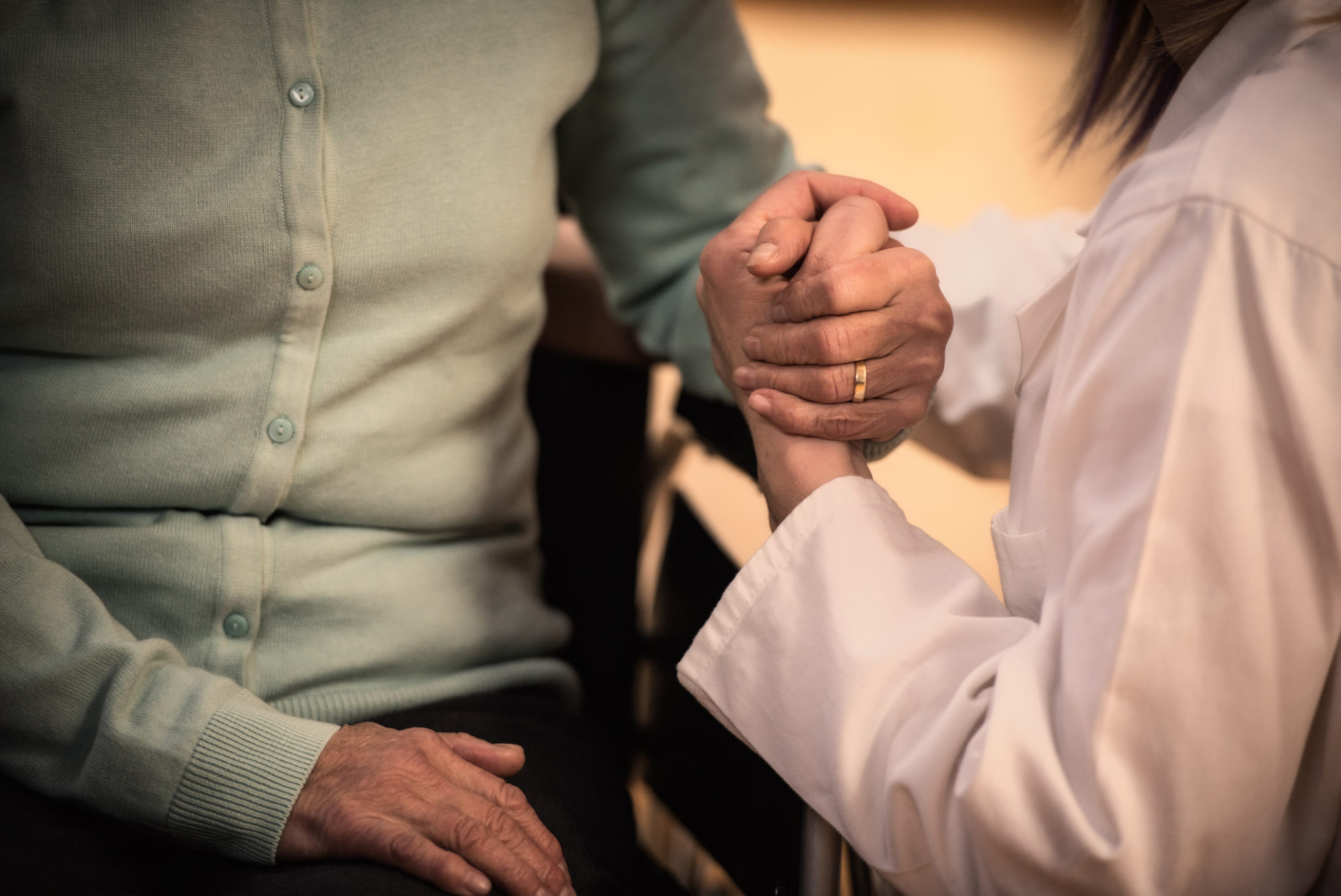 A nurse in a white coat tending to a disabled senior person, giving him a hand. Only torso and hands visible.