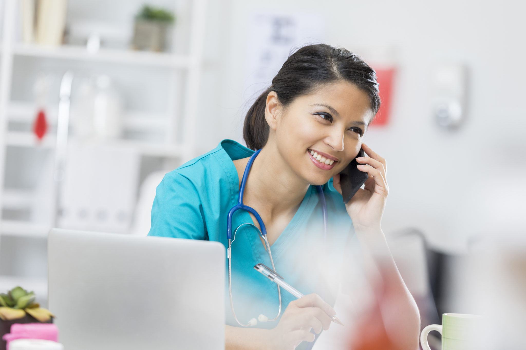 Smiling mid adult Asian nurse or doctor is on the phone with a patient. An open laptop is on the desk.