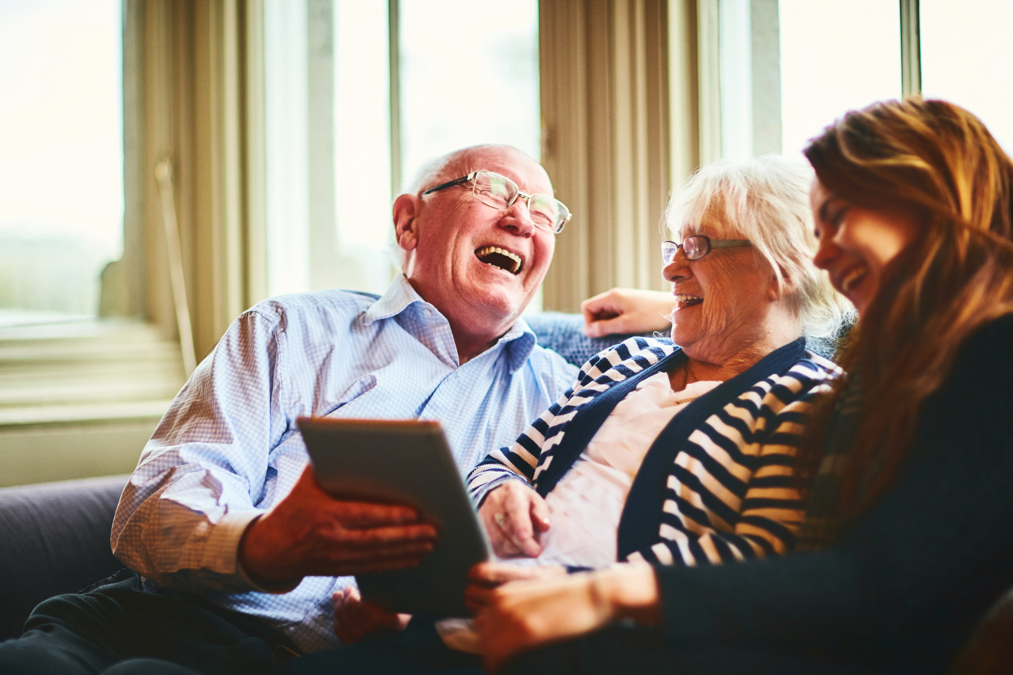 Cheerful senior man with his wife and daughter sitting on sofa at home with digital tablet