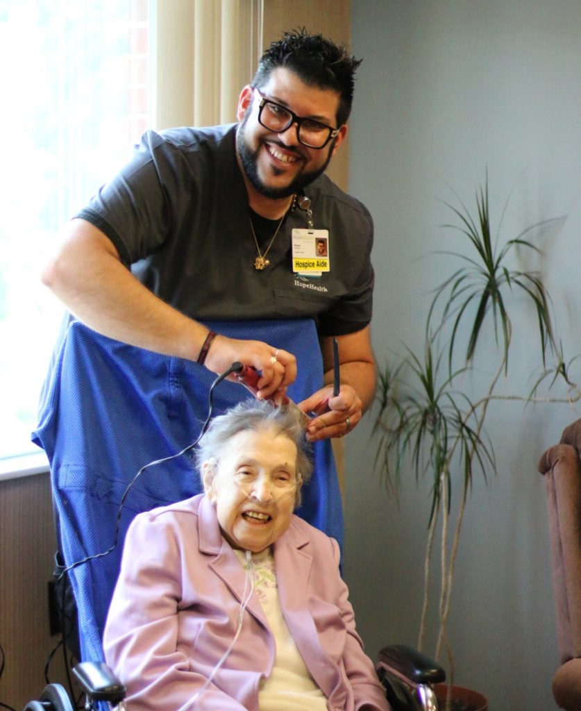 Man in grey scrubs stands over elderly woman in wheelchair while curling her grey hair.