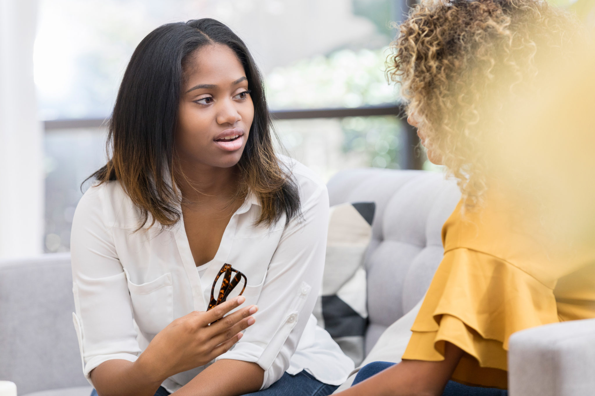 A young woman is serious as she holds a pair of eyeglasses and sits on a living room couch with an unrecognizable friend. She is asking her advice on a matter of concern.