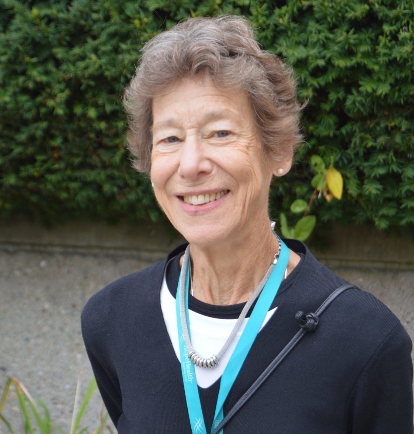 Older woman smiling and wearing a black sweater standing in front of a bush