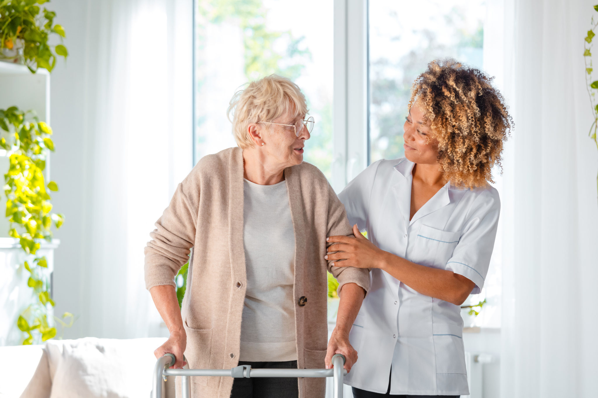 Afro american female home nurse supporting senior woman in using mobility walker at home.