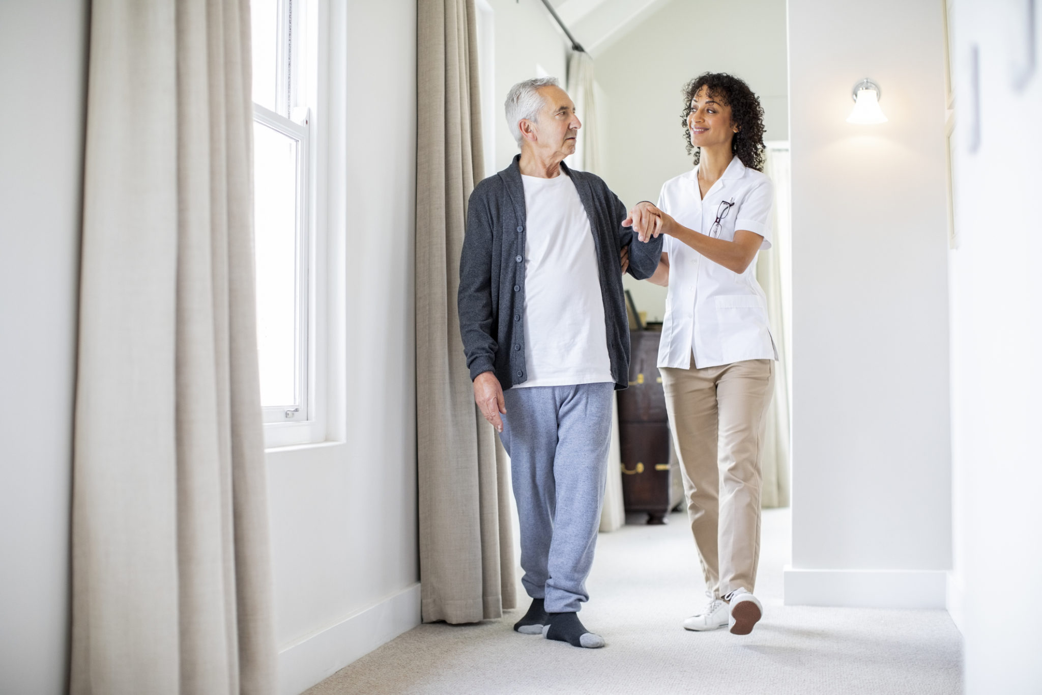 Full length of female caregiver walking with senior male patient. Retired man and nurse are in corridor at home. They are holding hands.