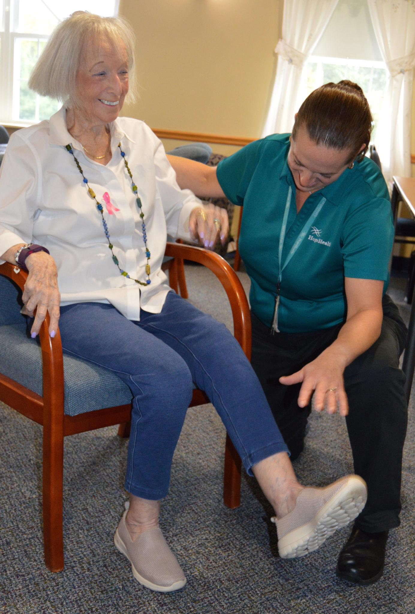 Elderly woman sitting in chair doing physical therapy exercises as a younger physical therapist is bending down to guide her through the exercises