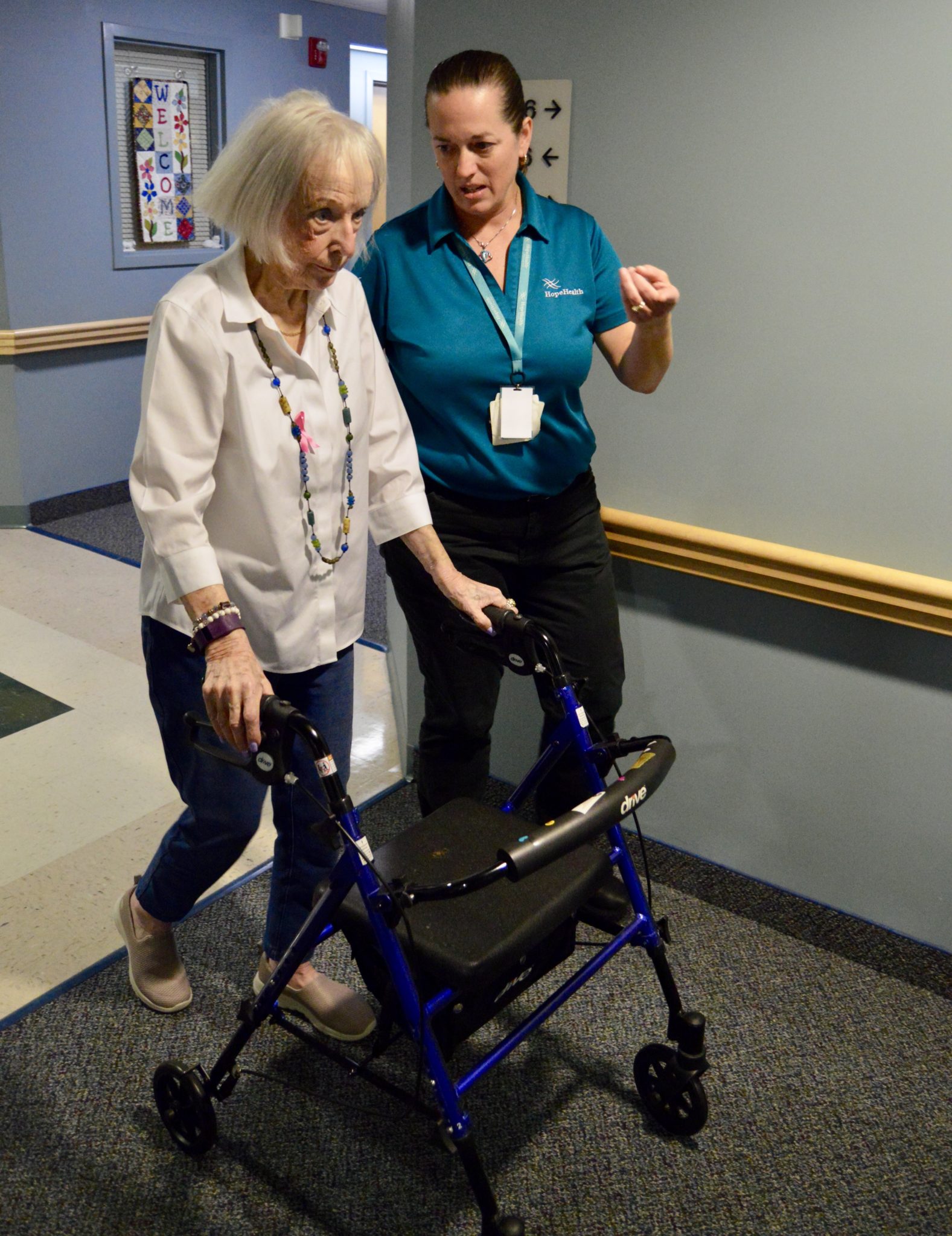 Elderly woman pushing a walker while a young female physical therapist guides her through the hallway