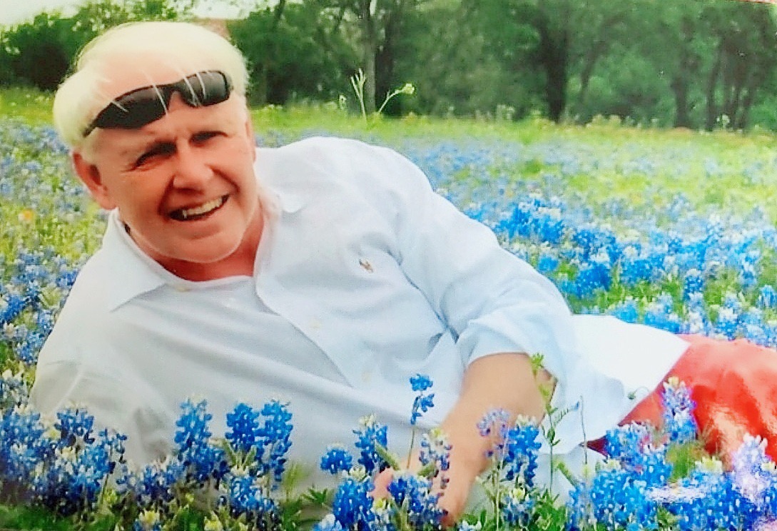 A man with white hair and a dress shirt with sunglasses on his head lays in a field of blue bonnets.