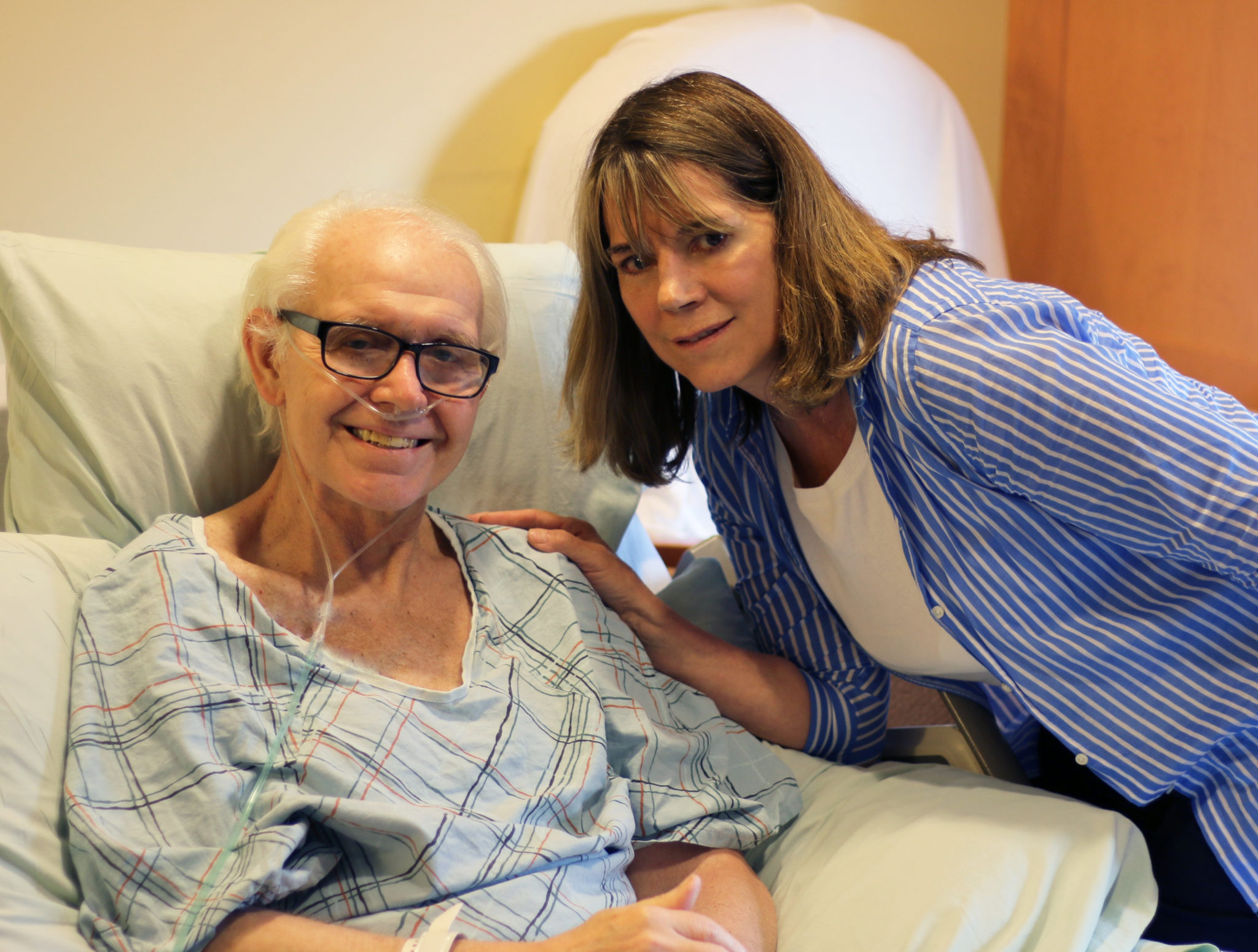 A man on hospice services wearing a hospital gown, in the inpatient unit smiles about his hospice experience, next to his supportive wife who leans close to him.