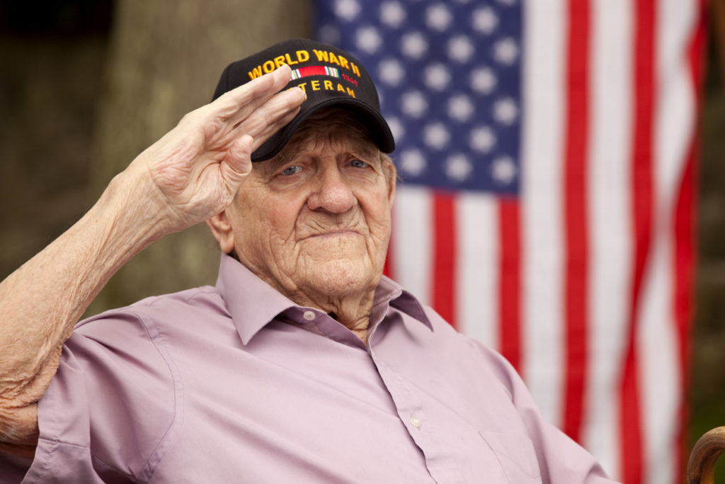 World War Two, Veteran wearing cap with text, "World War Two Veteran". Saluting Hand salute and looking directly in to camera with concerned look on his face, with US flag in background. Image shot with Canon 5D Mark2, EF 70-200 f2.8L USM lens, natural light.