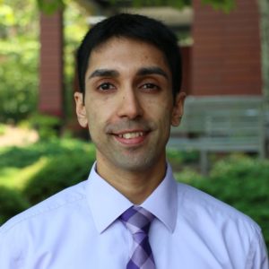 Man wearing purple business shirt and tie smiling in front of greenery