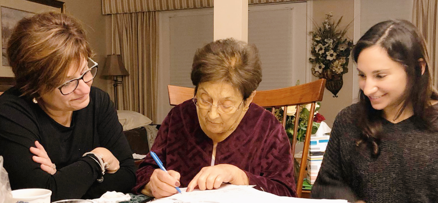 Three generations of women looking over a notebook as the grandma writes in her recipe book