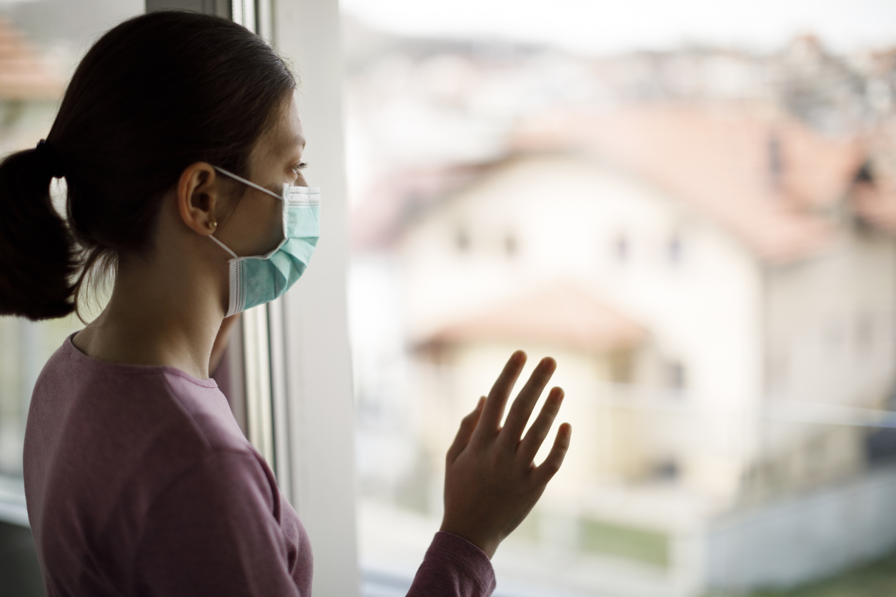 Portrait of sad teenage girl with face protective mask looking through the window at home grieving during coronavirus COVID-19