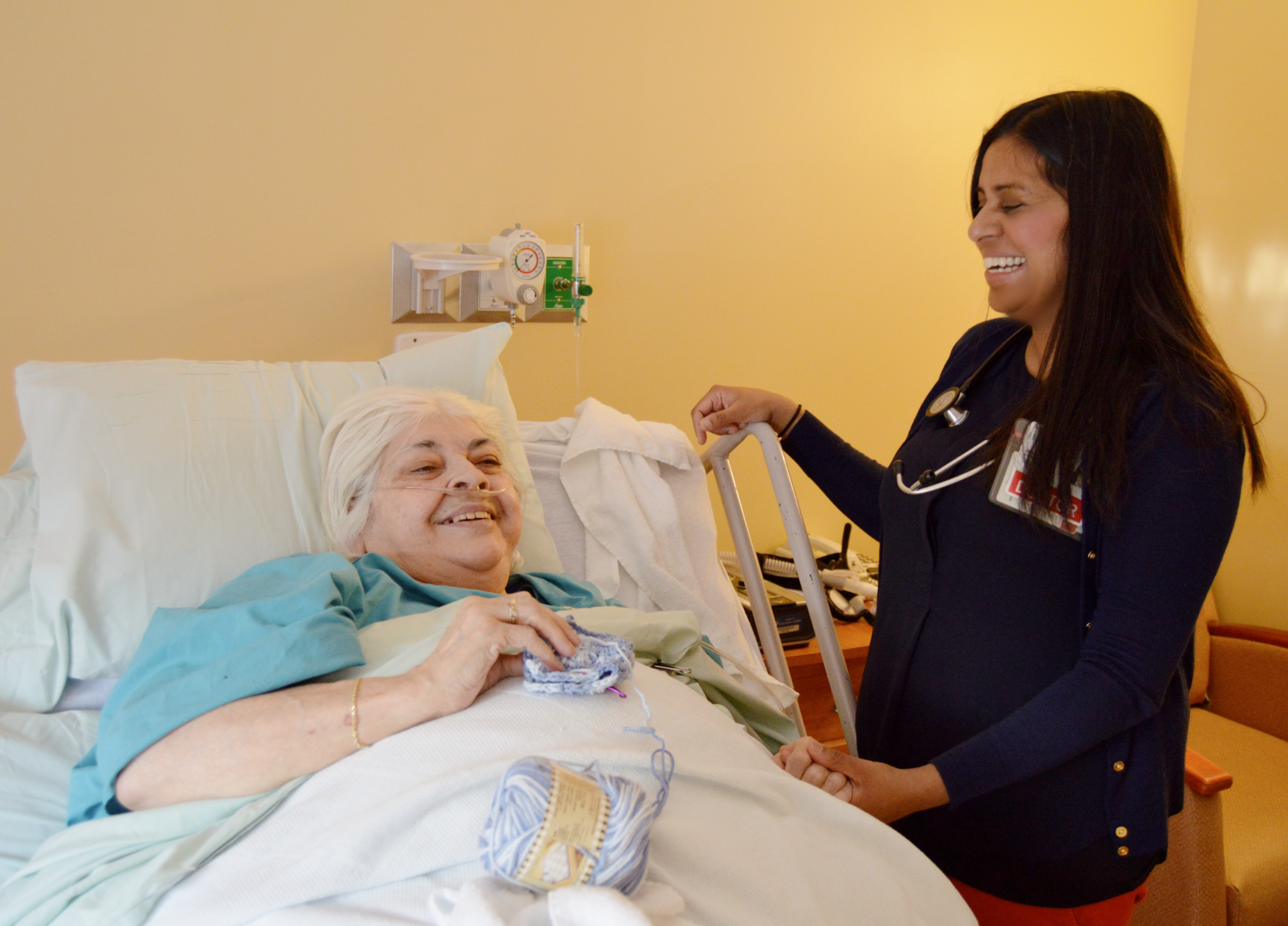 A woman laying in bed and knitting while she laughs and talks to her hospice doctor. She is receiving inpatient hospice care at the Hulitar Hospice Care Center