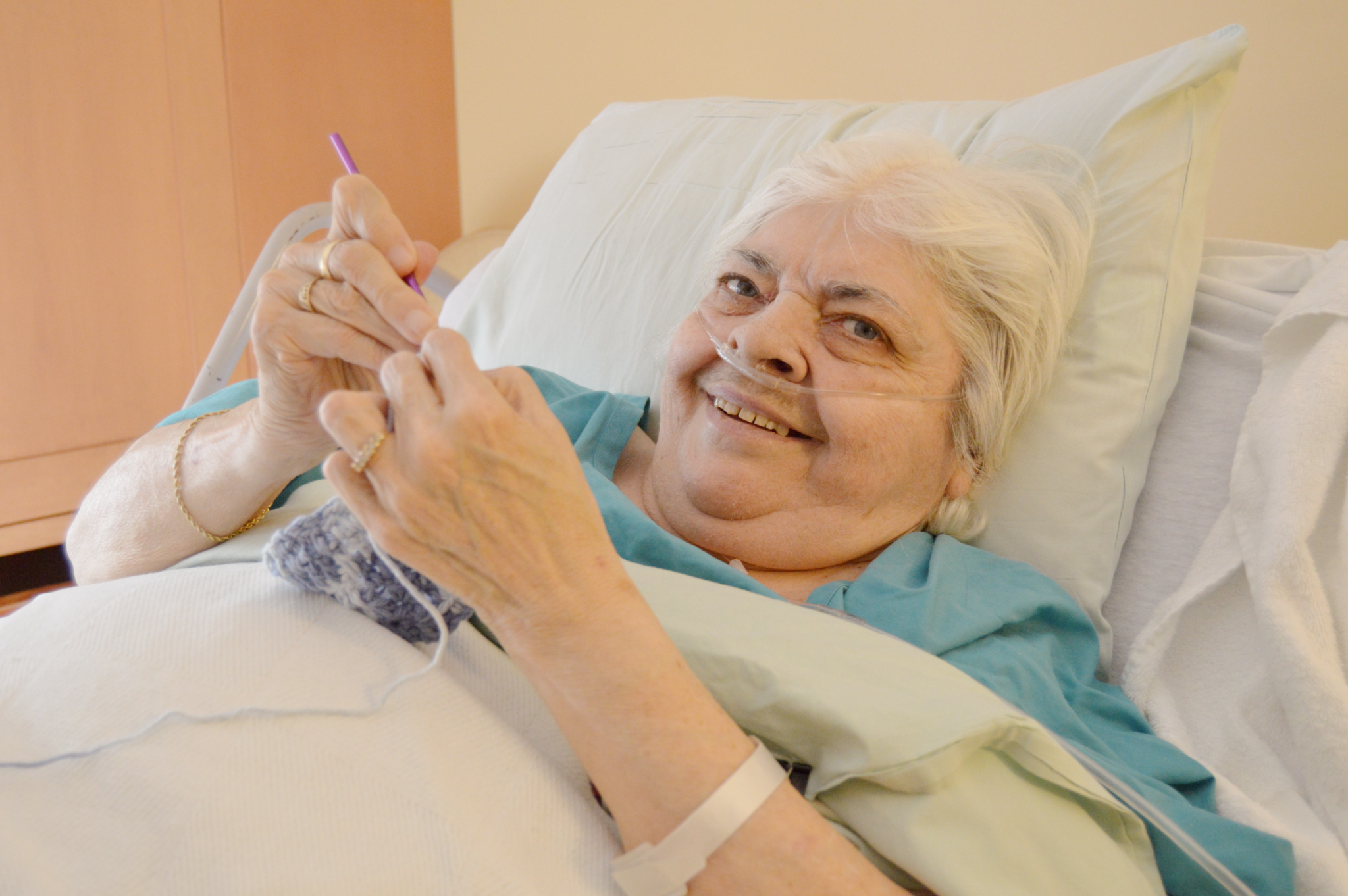 Woman laying in hospice bed knitting and smiling; through hospice care, she is able to stay comfortable at end of life