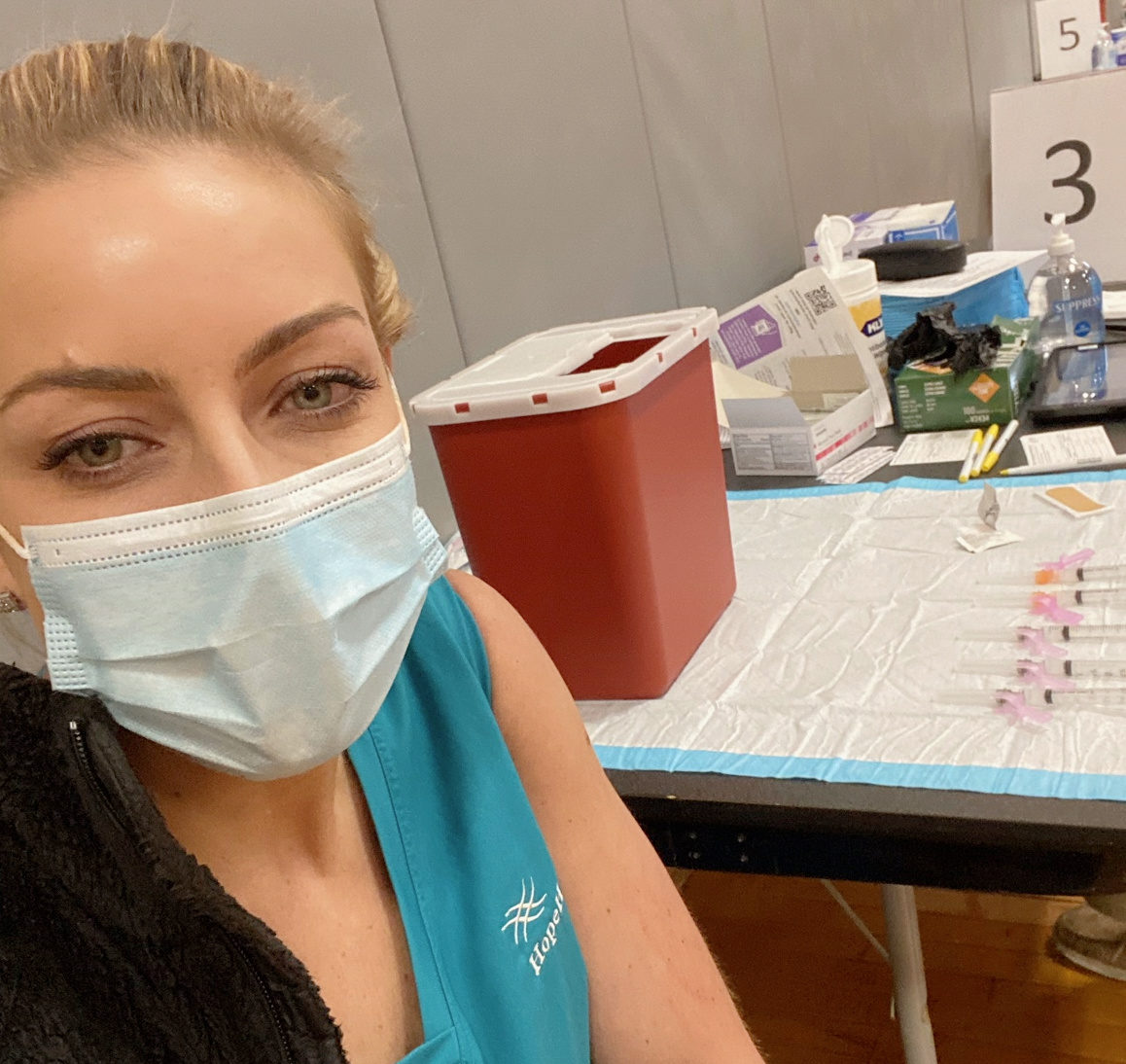 Female nurse wearing teal scrubs smiles as gets COVID-19 vaccine