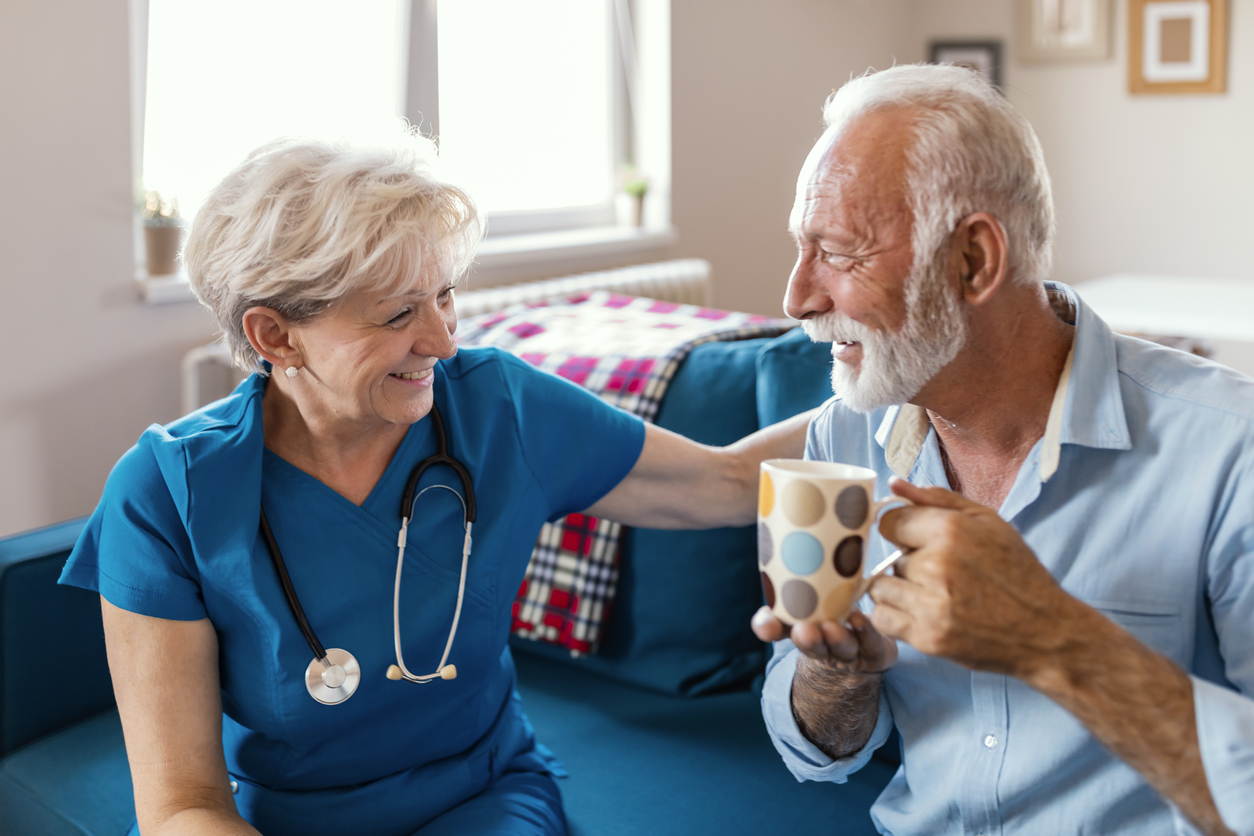 Float hospice nurse smiling at gray-haired male patient drinking from a mug.