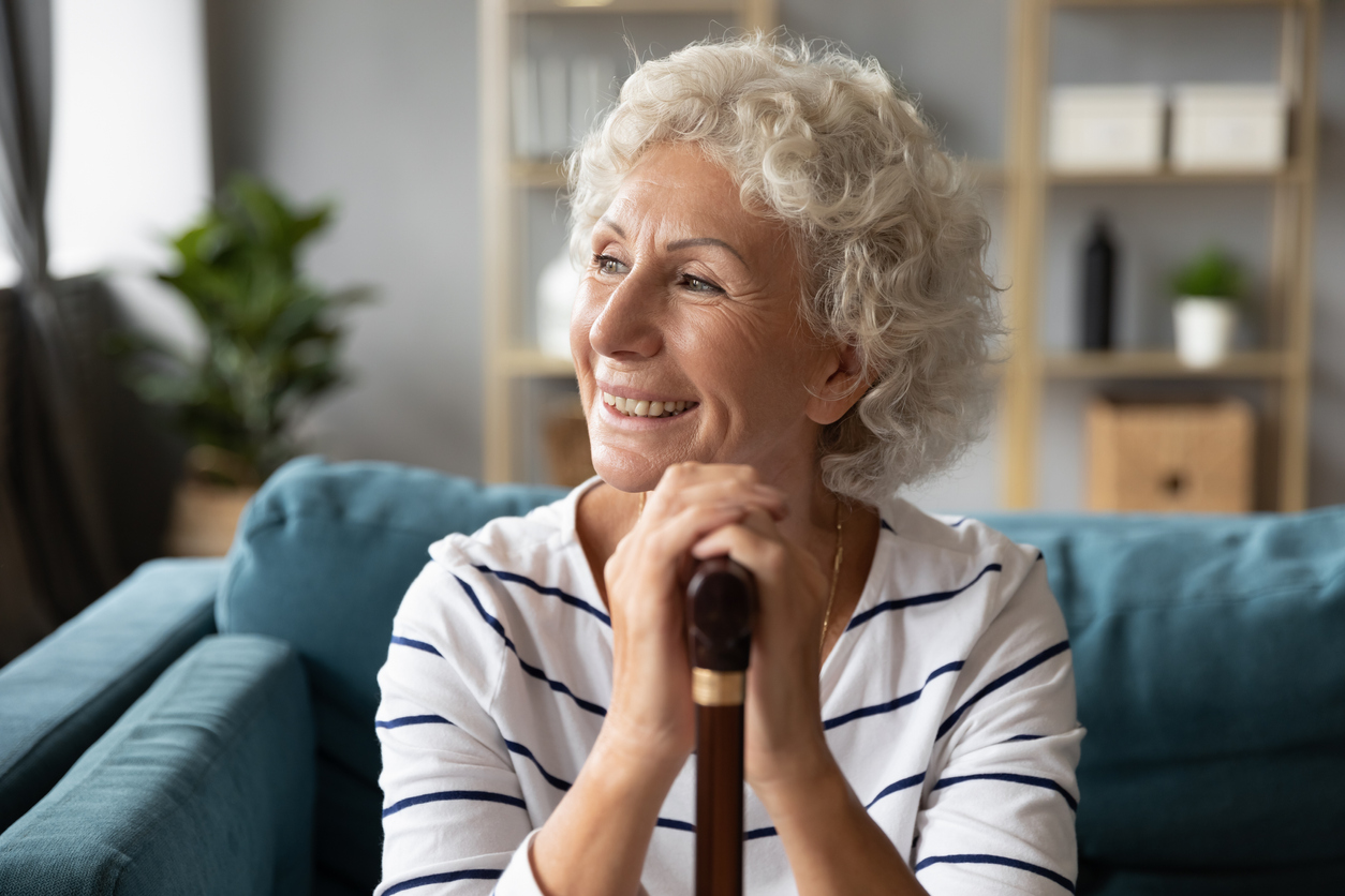 Happy smiling older woman holding hands on wooden cane, looking into distance, in window, dreaming, excited mature female using walking stick, sitting on cozy sofa in living room, feeling positive