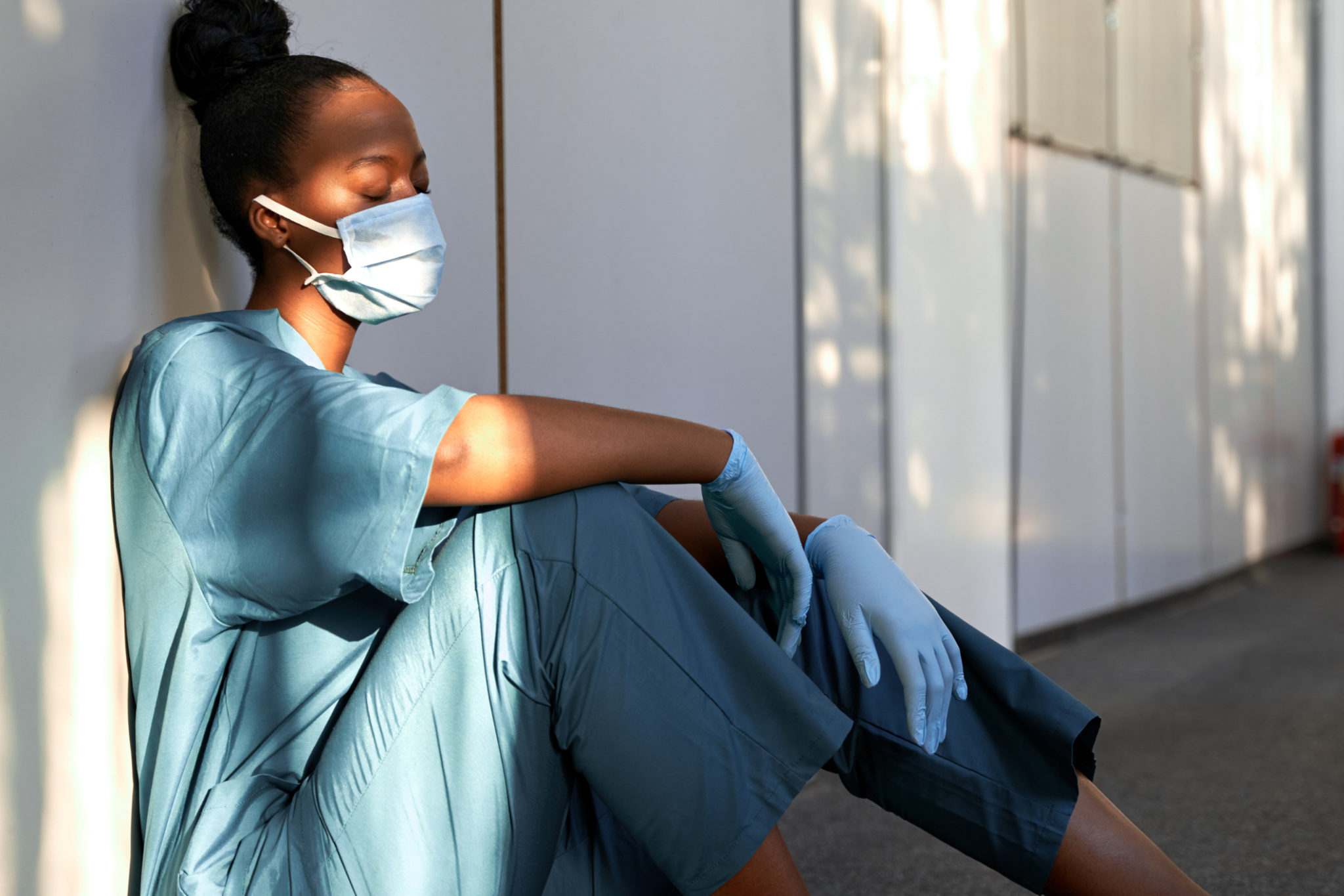 Tired exhausted female african scrub nurse wears face mask blue uniform gloves sits on hospital floor. Depressed sad black ethic doctor feels fatigue burnout stress, lack of sleep, napping at work. Tired exhausted female african scrub nurse wears face mask blue uniform gloves sits on hospital floor. Depressed sad black ethic doctor feels fatigue burnout stress, lack of sleep, napping at work.