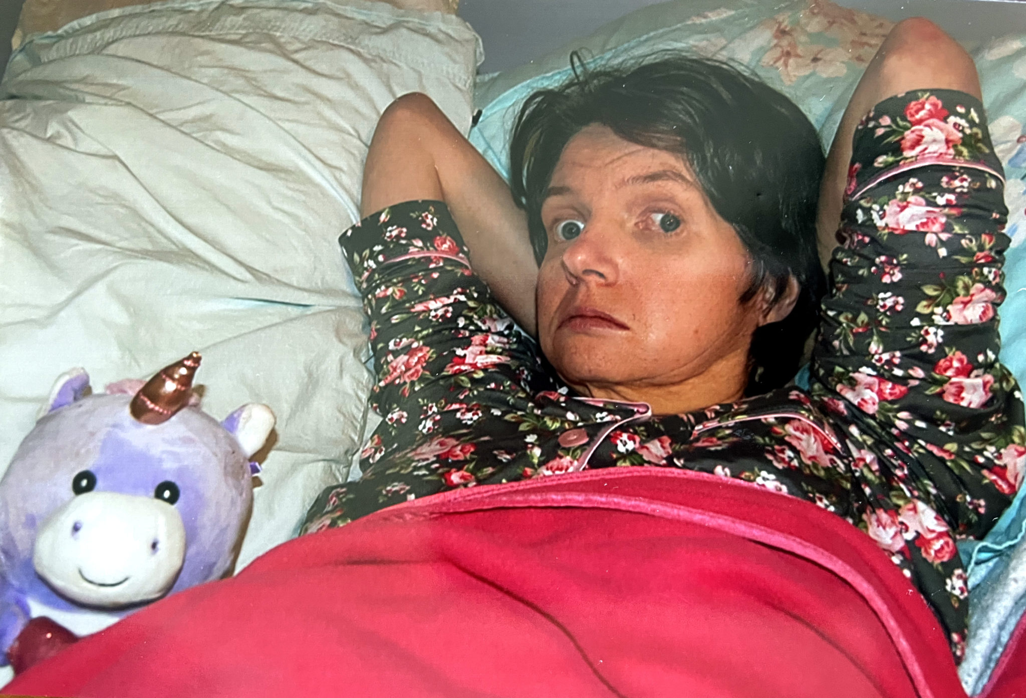 Woman in dark floral pajamas is tucked into bed with purple stuffed animal unicorn.