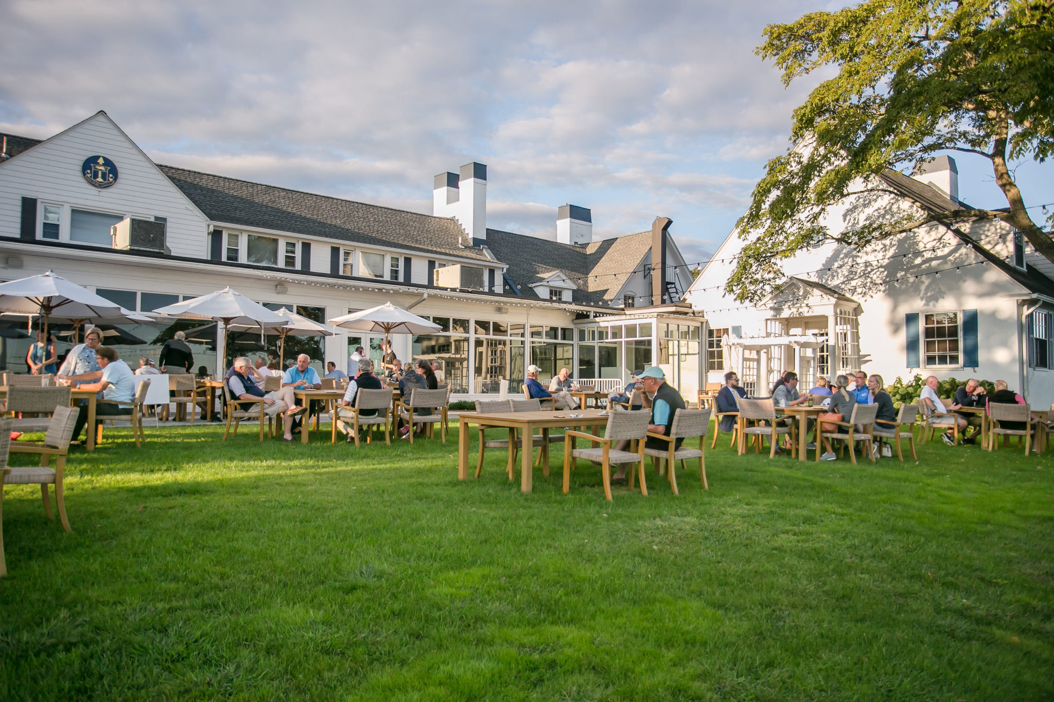 Large grassy area with people gathering for a golf invitational in front of a white building