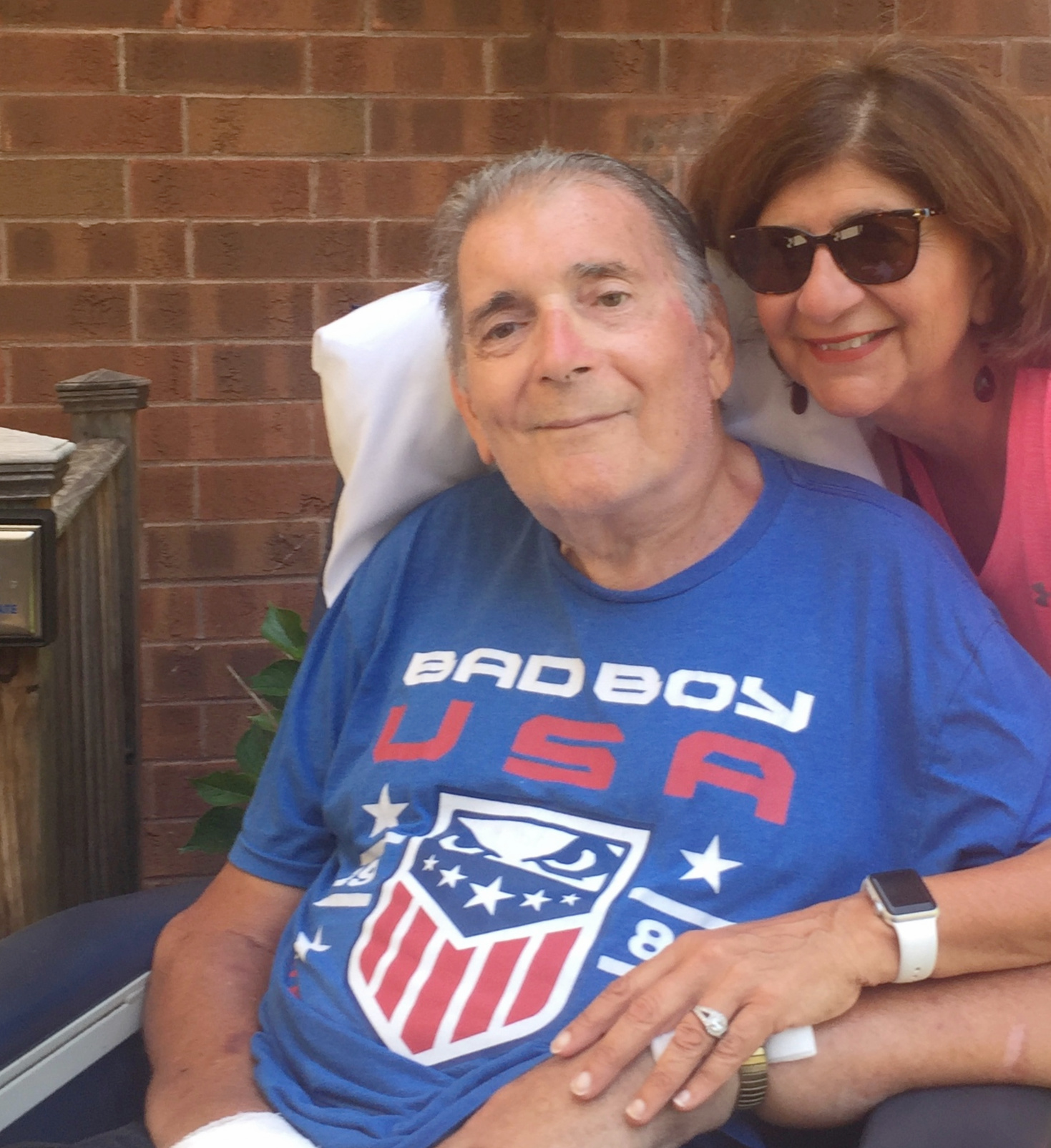 Senior man sitting in a chair with a woman behind his right side wrapping her arm around him