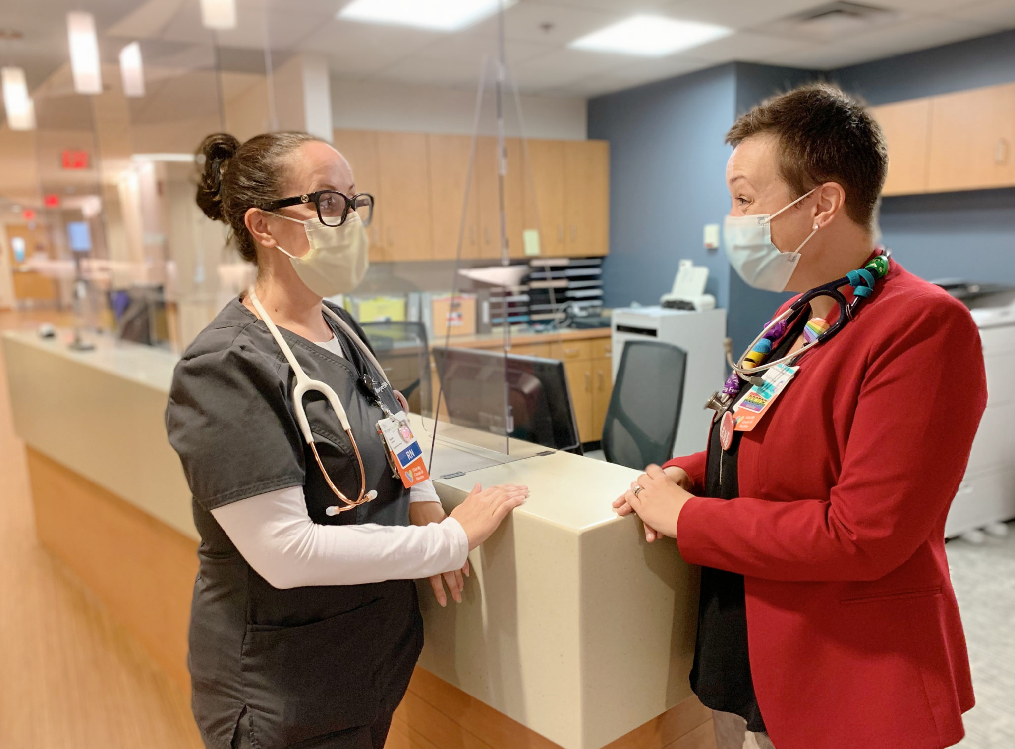 A nurse wearing grey scrubs and a surgical mask chats with a doctor wearing a red blazer and a surgical mask at the Hulitar Hospice Center