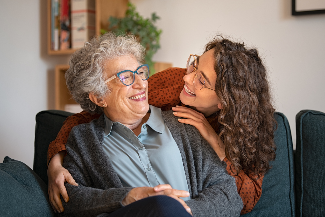 Grandmother and granddaughter laughing and embracing at home Old grandmother and adult granddaughter hugging at home and looking at each other. Happy senior mother and young daughter embracing with love on sofa. Happy young woman hugging from behind grandma with love.